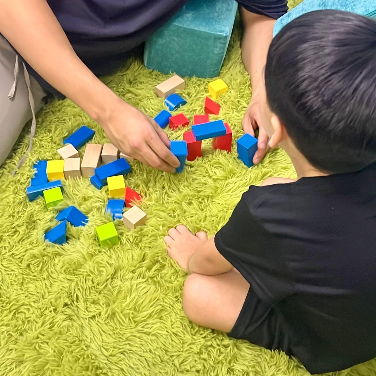 Un adulto y un niño arman juntos una torre con bloques de colores sobre una alfombra verde